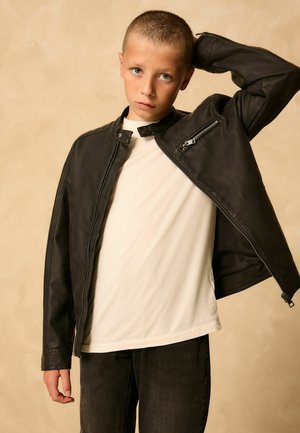 Jongen met kort haar draagt een zwarte leren jas over een wit overhemd, staand tegen een beige achtergrond met één hand achter het hoofd.