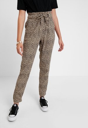 Vrouw draagt beige hoge taillebroek met luipaardprint en ceintuur, zwart T-shirt, schelpenarmband en zwart-witte sneakers.