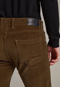 Brown corduroy pants with textured fabric, featuring a black leather label at the back waist and contrast stitching on pockets.