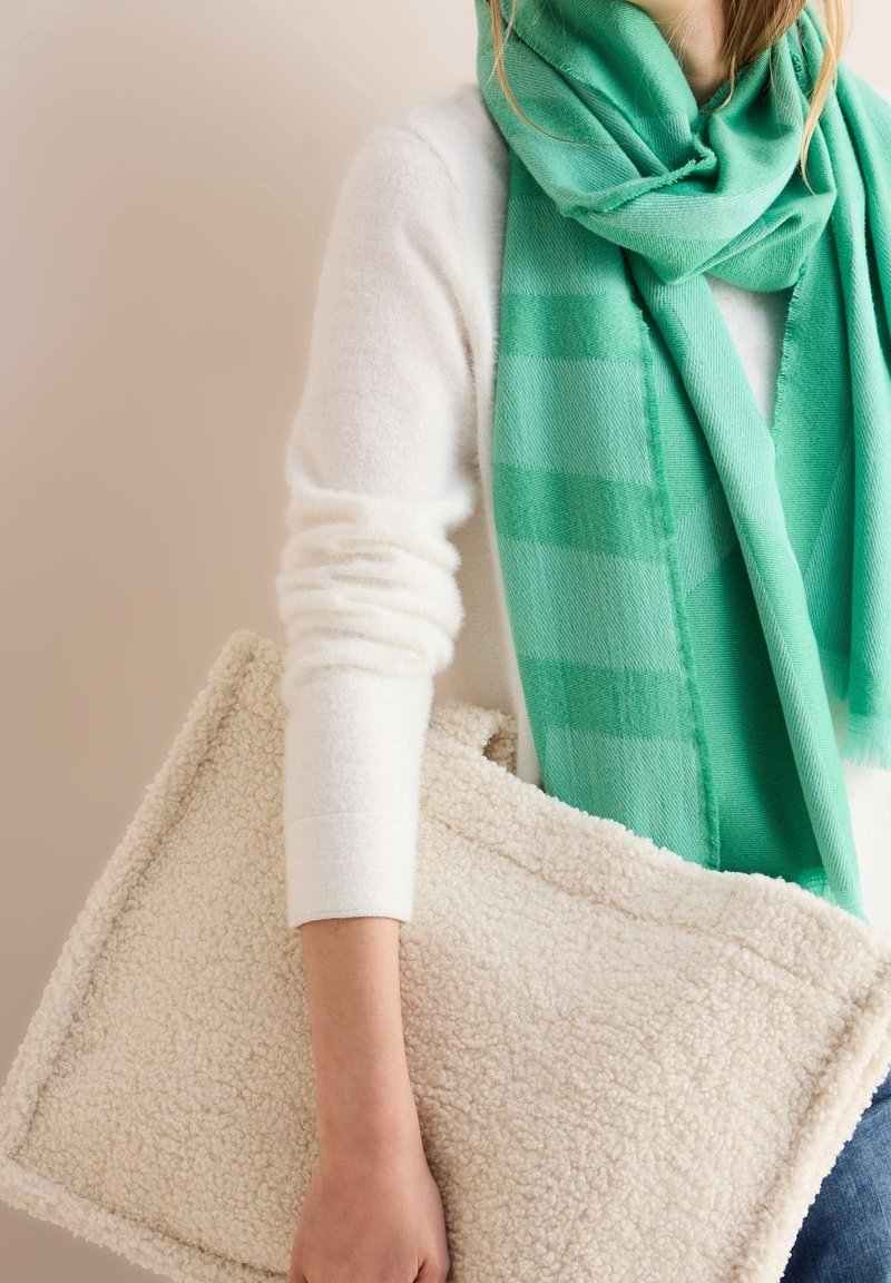 Personne portant un pull blanc et une écharpe verte tenant un grand sac fourre-tout rectangulaire en sherpa couleur crème sur un fond neutre.