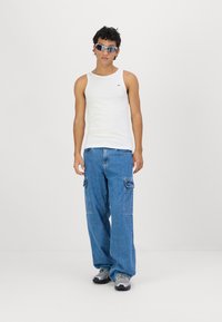 Fehér ujjatlan trikó, kék, bővös cargo farmernadrág oldalsó zsebekkel, szürke sportcipő és oversized napszemüveg. Minimalista háttér.