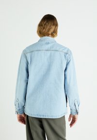 Veste en denim bleu clair avec une coupe décontractée, poignets boutonnés et col. Présente un ourlet droit et des détails de couture subtils.