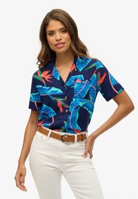 Korte mouwen marineblauw shirt met levendige tropische bladeren en bloemen. Heeft een knoopsluiting aan de voorkant en een borstzak, gecombineerd met witte broeken en een tan riem.