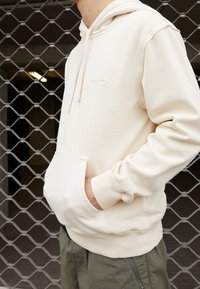Sudadera beige hecha de tejido suave, con un bolsillo frontal, capucha con cordón y puños acanalados. Detalle de logo minimalista en el pecho.