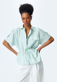 Mintgrünes, kurzärmeliges Blusenshirt mit Knopfverschluss, Brusttasche und verstellbarem Kordelzug in der Taille. Glattes, leichtes Material.