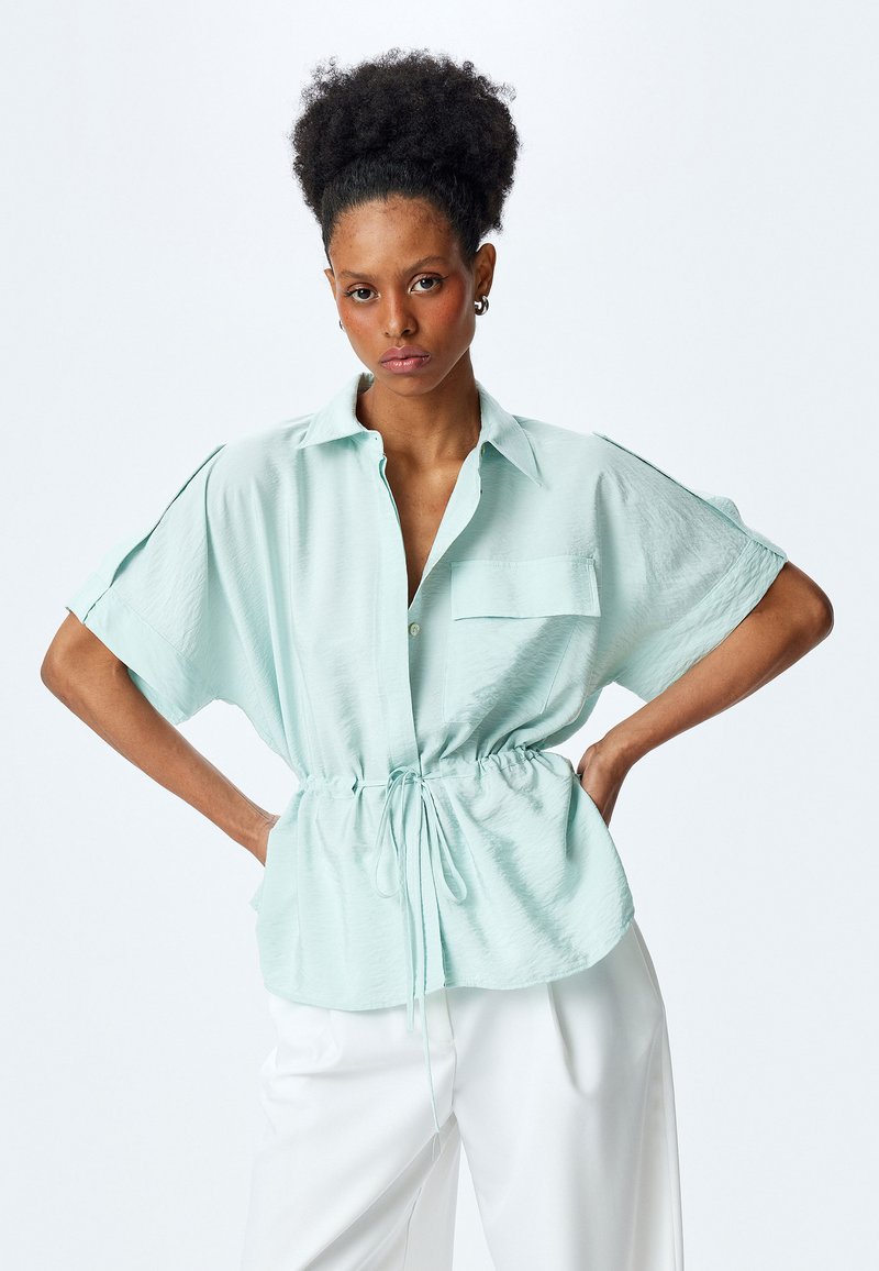 Mintgrünes, kurzärmeliges Blusenshirt mit Knopfverschluss, Brusttasche und verstellbarem Kordelzug in der Taille. Glattes, leichtes Material.