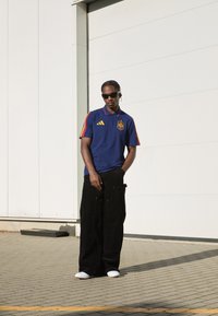 Zila polo krekla ar sarkaniem un dzelteniem akcentiem, apvienots ar platām melnām biksēm un baltām kurpēm. Tekstūras materiāli un sportiska dizaina elementi.