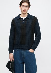 Gombos, nyakú kötött pulóver, váltakozó sötétkék és fekete textúrákkal. Három gombos záródás, bordázott mandzsetták és alsó rész, laza szabás.