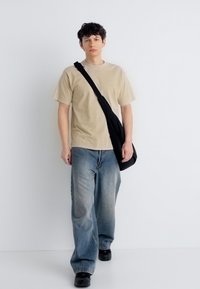 Νέος άνδρας που περπατά, φορώντας μπεζ t-shirt, φαρδιά μπλε τζιν, μαύρα παπούτσια και κρατώντας μια μαύρη τσάντα χιαστί σε λευκό φόντο.