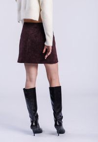 Mini-jupe en suède bordeaux associée à un pull crème ajusté ; le mannequin porte des bottes en cuir noir hautes au genou avec des talons aiguilles.