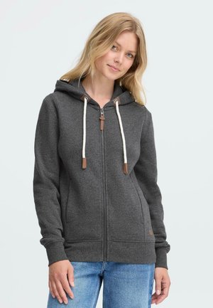 Femme blonde portant un sweat à capuche zippé gris foncé avec des cordons blancs et un jean bleu, se tenant contre un fond clair et uni.