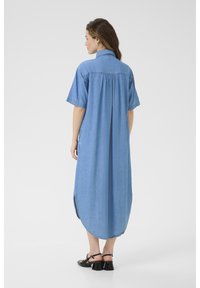 Svetlomodré denim šaty s krátkymi rukávmi, detailom rozparku na chrbte, bočnými rozparkami a zakriveným lemom. Nosí sa s čiernymi sandálmi na podpätku.