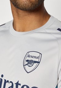 Grijze voetbalshirt met een marineblauw Arsenal-logo, ronde halslijn en korte mouwen. Gladde textuur met subtiele schouderdetails.