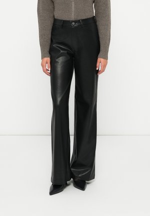 Vrouw in een zwarte hoge taille wijde broek van nep leer met een knoop en rits aan de voorkant, gecombineerd met puntige zwarte hakken en een bruine trui.