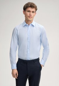 Helle blaue, langärmlige Hemdbluse mit Kragen und Knopfverschluss, aus glattem Stoff; kombiniert mit dunklen, maßgeschneiderten Hosen.