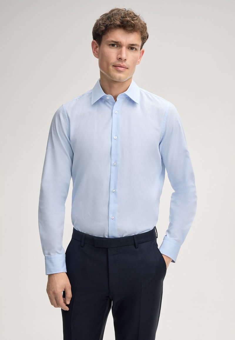 Camicia da uomo azzurro chiaro a maniche lunghe con colletto e chiusura a bottoni, realizzata in tessuto morbido; abbinata a pantaloni scuri e sartoriali.