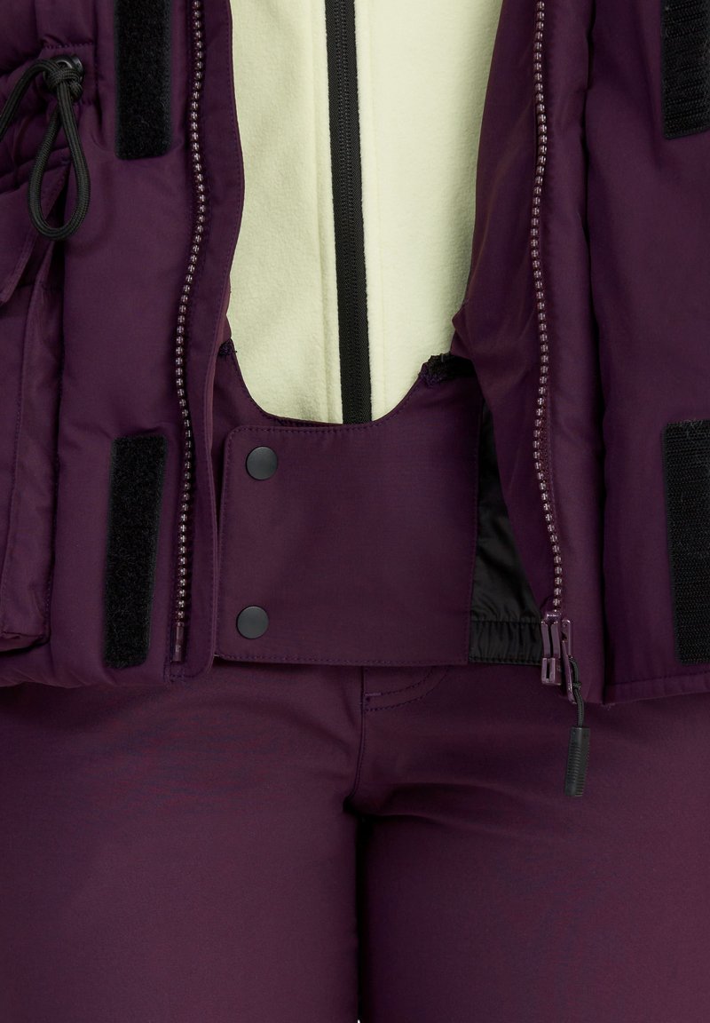 Lila isolierte Jacke mit Reißverschluss, Fleece-Futter, schwarzen Akzenten, Klettverschlüssen und Druckknopfverschluss. Passende, taillierte Hose ist unter der Jacke sichtbar.