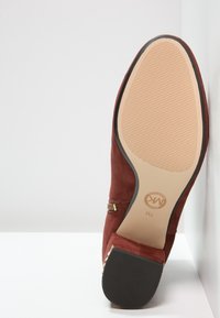 Chaussure à talon en suédine marron reposant sur le dos, montrant sa semelle beige texturée et son talon noir sur fond blanc.