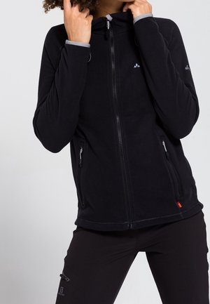 Personne portant une veste polaire noire zippée et un pantalon noir, ajustant le col de la veste avec les deux mains.