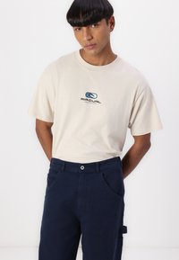 Beige cotton t-shirt with logo on the chest, paired with dark blue pants featuring a button closure and side pockets.