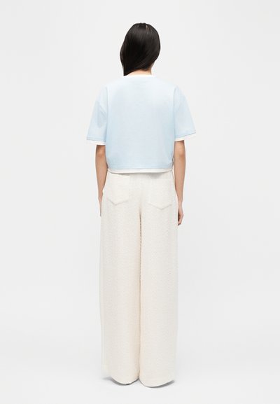 Γυναίκα με μακριά σκούρα μαλλιά που φοράει ένα κοντομάνικο ανοιχτό μπλε κοντό τοπ και παντελόνι κρεμ σε φαρδιά γραμμή με υφή, υψηλής μέσης, από πίσω.