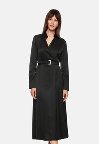 Zwarte satijnen jurk met lange mouwen, een V-hals, een ceintuur in de taille en een vloeiende rok. De riem heeft een metalen vierkante gesp als accent.