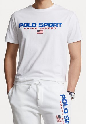 Bílý bavlněný tričko s výrazným modrým textem "POLO SPORT" a "RALPH LAUREN," s červeným akcentem a malou americkou vlajkou.