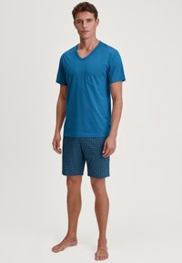 T-shirt en coton bleu à col en V avec une poche sur la poitrine, associé à un short marine à motifs avec des designs géométriques. Le mannequin est pieds nus.