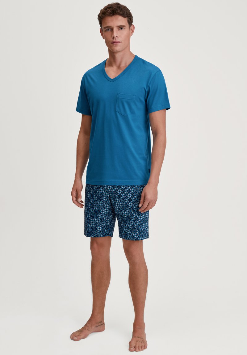 T-shirt en coton bleu à col en V avec une poche sur la poitrine, associé à un short marine à motifs avec des designs géométriques. Le mannequin est pieds nus.