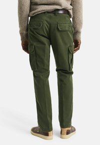 Mann in olivgrünen Cargo-Hosen mit Klappentaschen, braunem Gürtel, beigem Pullover und braunen sowie beigen Sneakers, der vor einem weißen Hintergrund steht.
