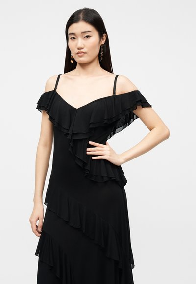 Νεαρή γυναίκα με μακριά σκούρα μαλλιά, φορώντας μαύρο φόρεμα με βολάν και έξω τους ώμους, στέκεται με το ένα χέρι στη μέση μπροστά σε απλό φόντο.