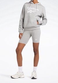 Felpa grigia Reebok con logo bianco, abbinata a pantaloni corti grigi. Si indossano sneakers bianche e calzini alla caviglia. Tessuto liscio, vestibilità casual.