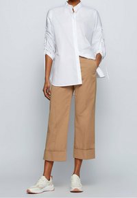 Witte button-up shirt met een hoge kraag en geplooide mouwen, gecombineerd met lichtbruine wijde broekspijpen en witte sneakers met roze accenten.