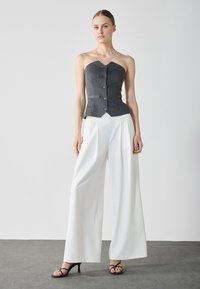 Top fără bretele, cu dungi gri și un design ajustat, închidere cu nasturi, purtat cu pantaloni largi albi și sandale cu toc negre, pe un podea gri.
