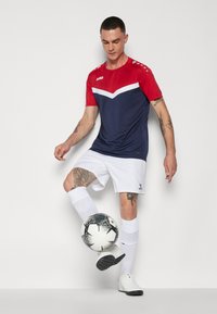Rode en marineblauwe voetbalshirt met korte mouwen met een chevronpatroon, gecombineerd met witte shorts en sokken. Houdt een zwart-witte voetbal vast.