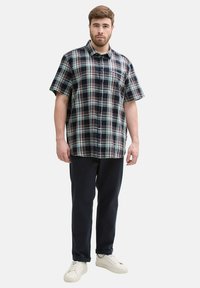 Camicia a maniche corte a quadri in navy, verde e rosso con colletto button-down, abbinata a pantaloni scuri e sneakers bianche.