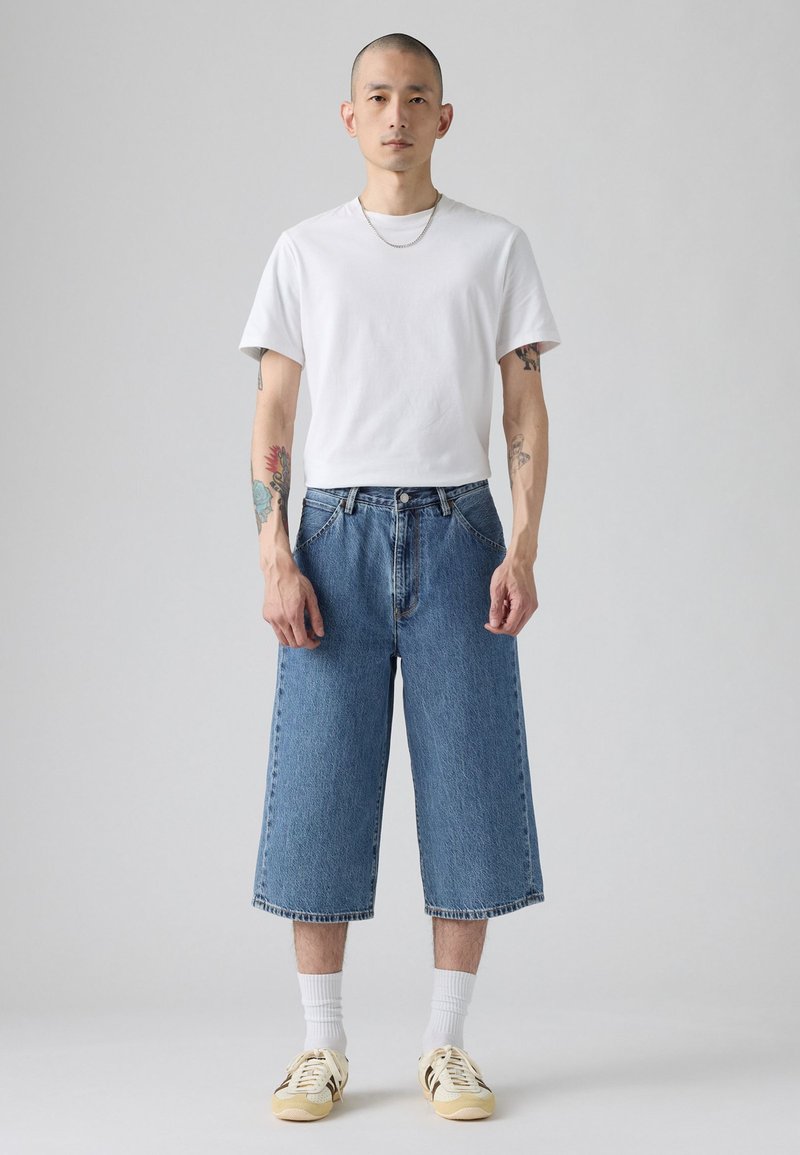 Culottes de mezclilla de color azul claro con diseño de corte recto, combinados con una camiseta blanca lisa y zapatillas beige. Presentan un cierre de botones a la vista.