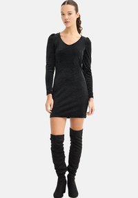 Vestido mini negro de manga larga con lunares blancos, con escote en V y diseño ajustado, combinado con botas negras hasta la rodilla.