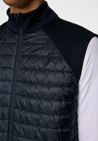 Mörk marinblå väst med kviltad textur och hög krage. Har slätt tyg på axlarna och dragkedjestängning. Logotyp på kragen.