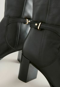 Botas de cuero negro hasta el tobillo con tacón cuadrado, un diseño elegante con una correa decorativa y acentos de hardware en tono dorado.