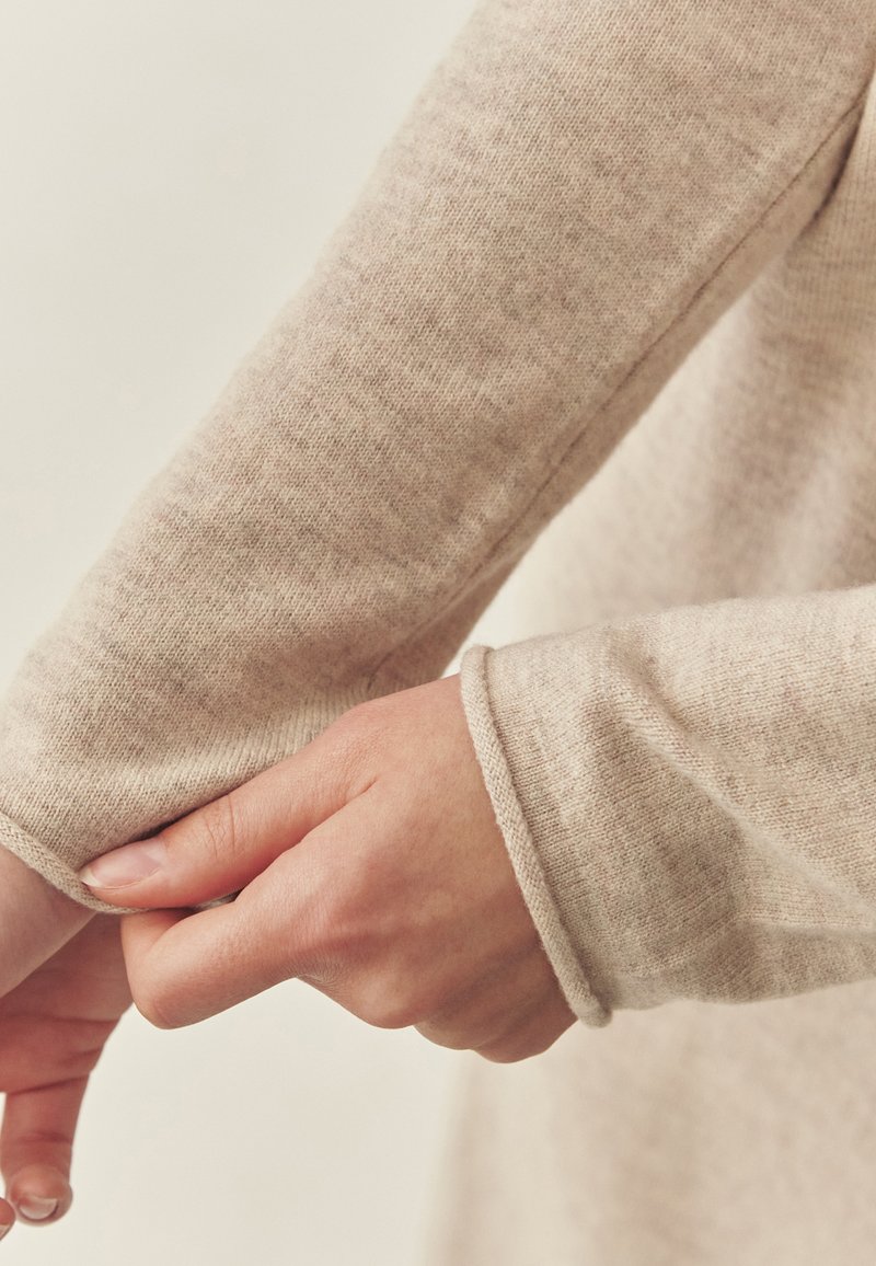 Jasno beżowy sweter z dzianiny z dopasowanym mankietem. Tkanina ma miękką teksturę, pokazując subtelny melanżowy wzór i staranne detale przeszycia.