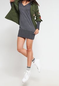 Graues Strickkleid mit V-Ausschnitt und geripptem Saum, kombiniert mit einer olivgrünen Jacke mit Applikationen, weißen High-Top-Sneakern und gestreiften Socken.