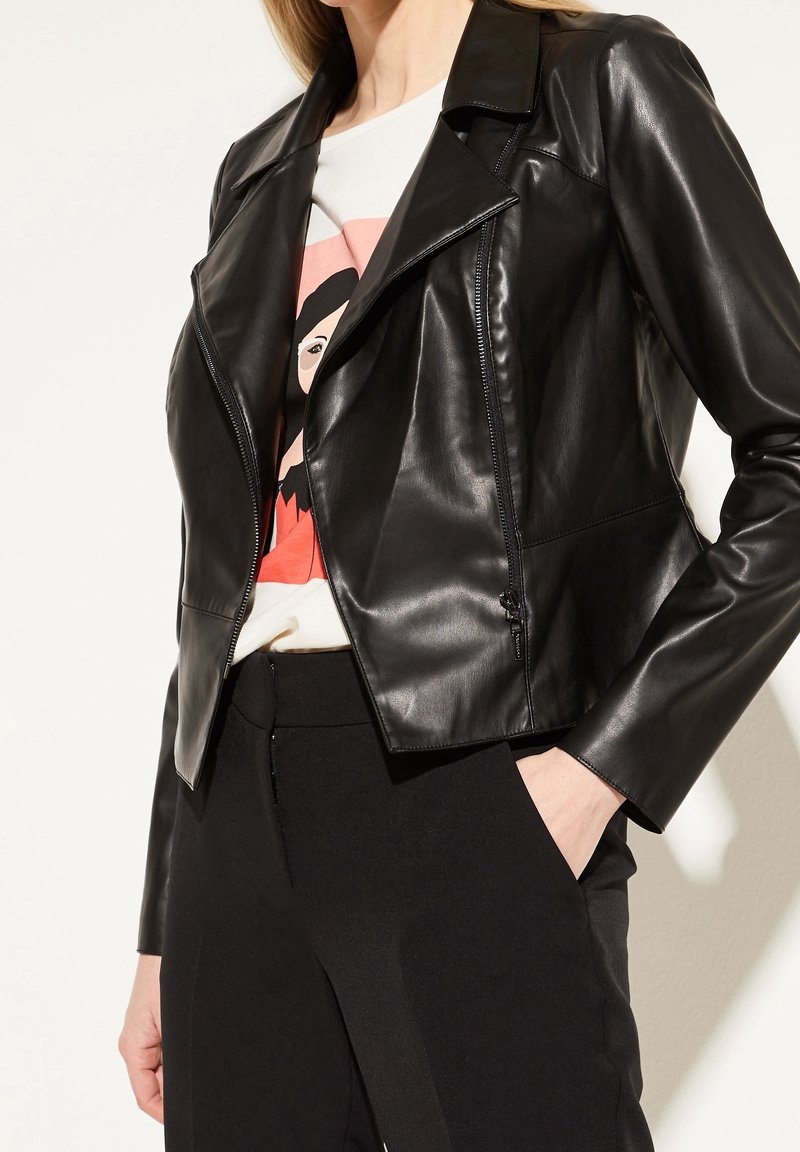 Veste en cuir noir au design ajusté, avec une fermeture éclair à l'avant et des revers larges, portée sur une chemise blanche ornée d'un graphisme coloré.