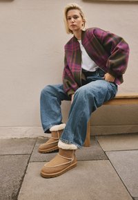 Bottines en daim marron avec doublure en fourrure, couture décorative prononcée et semelle texturée. Le modèle porte un jean bleu ample et une veste colorée.