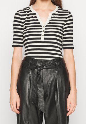Vrouw draagt een zwart-wit horizontaal gestreept T-shirt met korte mouwen, V-hals en knopen, gecombineerd met hoge zwart leren broek met een gestrikte riem.