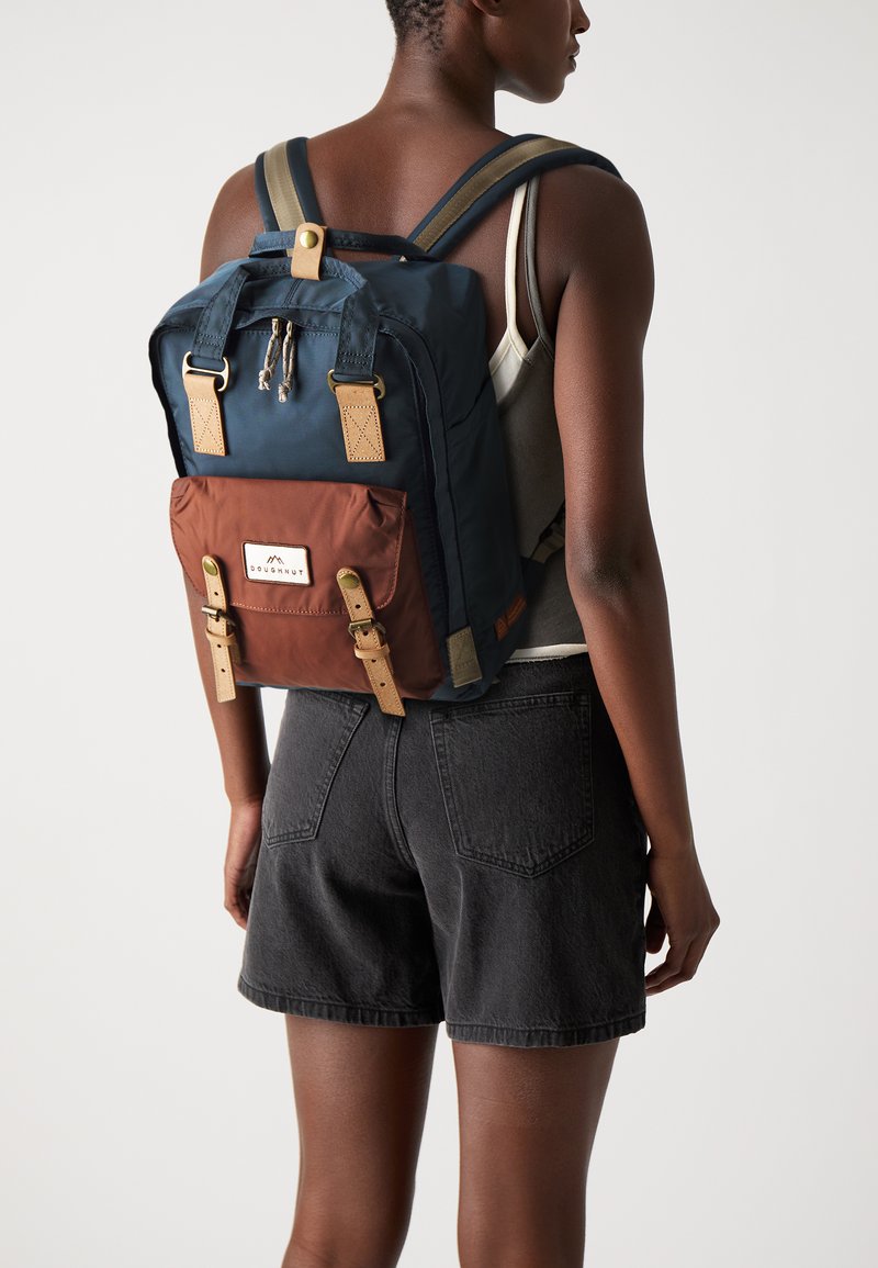Navy and brown backpack with gold hardware, leather accents, and adjustable straps; features a front pocket with a branded label.