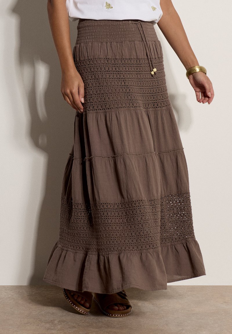 Jupe longue marron à étages avec des détails en crochet, portée avec un haut blanc et un bracelet doré, associée à des sandales foncées sur un sol neutre.