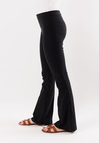 Pantaloni neri a zampa con vita alta, caratterizzati da una texture liscia e un materiale elasticizzato. Indossati con sandali marroni con listini.