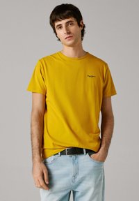 Jonge man die een mosterdgele Pepe Jeans T-shirt en lichtblauwe jeans met een zwarte riem draagt, poserend met één hand in de zak.