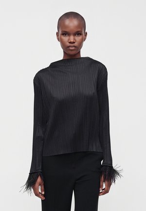 Femme aux cheveux courts portant un haut noir à manches longues avec de fines rayures verticales et des poignets à franges, associé à un pantalon noir.